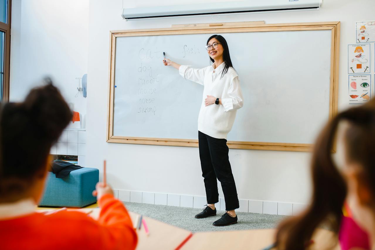 Na obrazku widać nauczycielkę, stojącą przed tablicą, trzymającą kredę i wyjaśniającą materiał uczniom